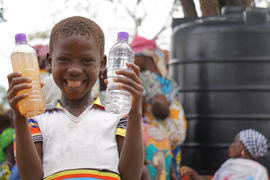 Photo of a young smiling girl holding two plastic water bottles, one with brown liquid inside, the other with clear liquid. A large black container and two women are behind her