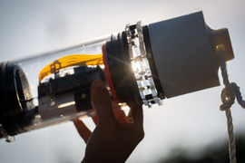 Hand holding up the microbe-monitoring system, which is a clear cylinder with electronics inside and a rope tied to the top