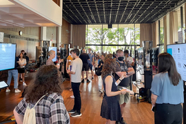 A photo, taken at the MIT Ethics Sustainability Student Showcase, shows attendees and students chatting with each other. Large TV screens show student projects at various stations.