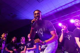 A diverse group of 8 MIT students, wearing black t-shirts and singing on a stage. The photo centers on a young man, who sings a solo.