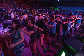 Photo of at least 100 people in a darkly lit dance hall, many of whom are passing in front of the camera in a conga line