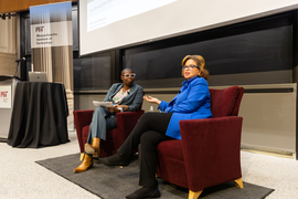 Harriel, left, listens as Jarrett speaks in front of the audience. They are sitting on red chairs and a large blackboard is in the background.