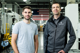 Martin Feldmann in a gray t shirt and John Hart in a black sweater vest face the camera in front of industrial equipment