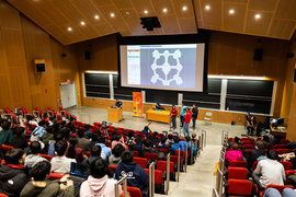 Projected screen at front of room shows a virtual battlefield. There are seven individuals on stage and three seated at the side while group of people seated in auditorium seats looks at the screen.