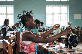 A woman with a mohawk-style braided hairstyle works at a sewing machine with brightly-colored fabrics, alongside other women 