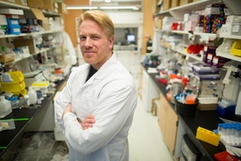 Dan Anderson stands in his lab, wearing a lab coat. The lab is full of equipment on tabletops and shelves.