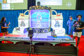 Avani Narula and Amir White stand on opposite sides of the game board, each giving a thumbs up while controlling their robot on the board. The course includes rocks, ramps, gears, and red balls. In the center is a unique DNA helix made of white balls.