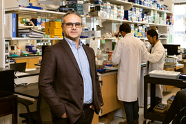 Yilmaz stands his lab, filled with various lab equipment, including binders and boxed supplies. 2 people in lab coats are in the background working together at a table.