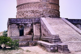 The image highlights a wide staircase leading up to a cylindrical structure. The building is weathered and some foliage is in the foreground.