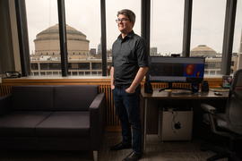 McDonald stands in his office, with a sofa and computer next to him. Through the large windows is MIT’s Great Dome on an overcast day.