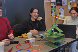 3 students smile at a table. A gyrating green structure as a small hula-hoop spinning around it.