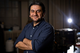 Tasan smiles with arms crossed in dark room, with spotlight shining in background on right.