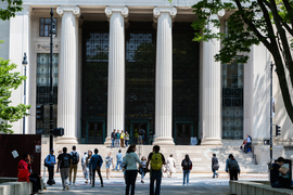 People cross Mass. Ave intersection, with a view of the steps and columns of Lobby 7, on a sunny day.