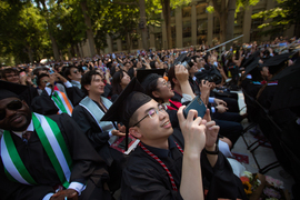 The seated graduates, wearing caps and gowns, use their cameras to take photos of the flying drone, not seen.