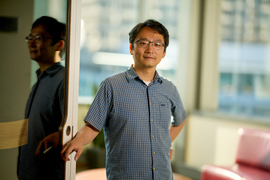 Andy Sun poses for portrait during the daytime next to reflective door while resting their right hand on handle. The blurry background has glass window with view of a building, a red lounge chair, and a round table.