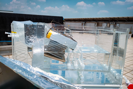 A desalinization prototype setup, a clear rectangular box with waters, tubes, and square spring surrounded by a larger rectangular box, outside in the sun.