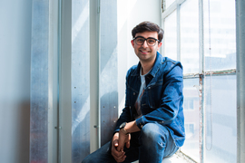 Adi Mehrotra smiles while sitting on a window ledge, with bright light streaming in from the window.