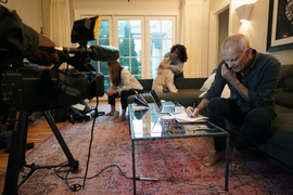 Moungi Bawendi sits on a sofa in his living room, looking at laptop in front of him on a coffee table and speaking into a cell phone. Family members are in the background; a large camera on a tripod is in the foreground.