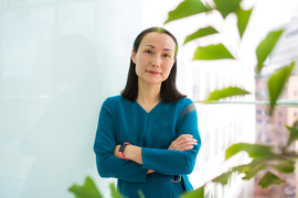 Haruko Wainwright portrait with arms crossed against a bright white background, with blurry plant leaves in foreground.