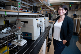 Desirée Plata stands next to a table in a lab full of equipment. On the table is a bubbling glass beaker and 3 machines labeled “Thermo Scientific.”