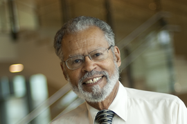 Portrait photo of Willard Johnson, who stands indoors with shiny items on a wall behind him out of focus