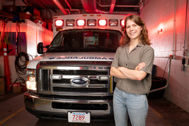 Abigail Schipper smiles with arms crossed while standing in front of a parked MIT ambulance with red lights on.