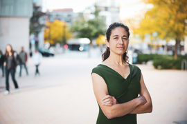 Elsa Olivetti stands outside with arms crossed. In the blurry background are yellow trees and people walking.