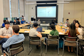 A meeting room is filled with about 20 students, seated, in one-on-one conversations with each other. A projection screen in the background shows a document with the title “Who Are You?”