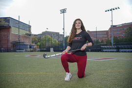 Liberty Ladd wears an MIT field hockey shirt and kneels on a field hockey field, holding a field hockey stick.