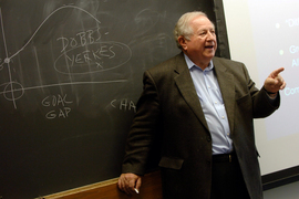 Ed Roberts stands with his back to a chalkboard with graphs and notes written on it. He is speaking and pointing with one finger.