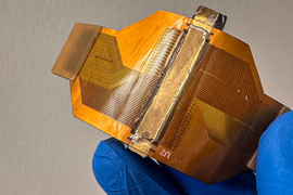 A blue-gloved hand flexes a small orange wearable sensor, with conductive traces on the circuit board.