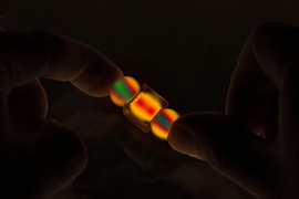 In darkness, thumbs hold two small spherical and one small cube objects that light up in vivid yellow, red, and green.