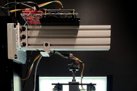 In the lab, the prototype scanner is made of metal and wires, with a sturdy clear box inside. Inside the box is a glass tube with yellow material inside. A lightbox is in the background, providing light.