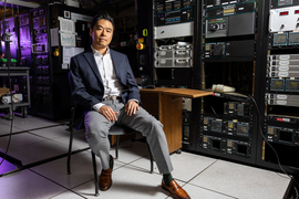 Jeehwan Kim sits on a chair in a dark lab filled with equipment, with a purple light in background.