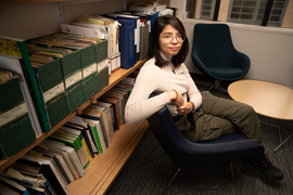 Taken slightly from above, photo shows Nicole McGaa sits on a dark chair in an office with shelves filled with papers and books.