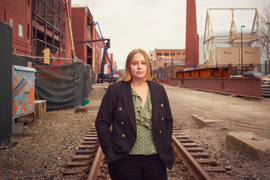 Mikayla Britsch stands on railway tracks near MIT campus, with buildings in background.