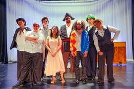 Eight people in costumes pose while on stage. Some are dressed like pirates, clowns, and some wear vintage clothing.