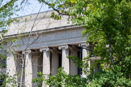 The columns of Building 7 are visible through green foliage on a sunny Spring day.