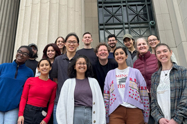 About 15 class participants smile for a group photo in front of MIT columns.