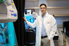 Sebastian Lourido wears a lab coat with his name, and stands in a lab with blue-lit equipment.