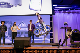 A child mannequin is on a bike, which is tilted on a machine, as four students on stage talk about their design.