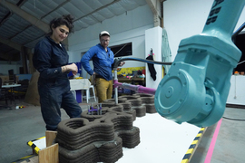 Two people watch a robot arm print a 3D cement wall.