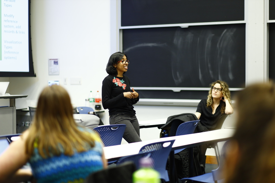 Basuhi Ravi lectures in front of a classroom, leaning against a table, her hands clasped in front of her.